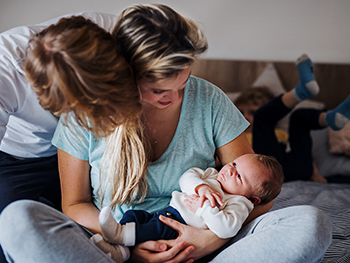 Parents sitting on bed with newborn baby and toddler in the background.